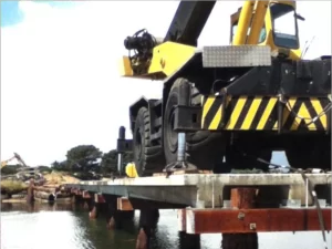 A large yellow crane is precariously positioned on the narrow, unfinished Barnbougle Bridge over water, with some wheels near the edge. The bridge is supported by wooden and metal pilings, with trees and construction material in the background.
