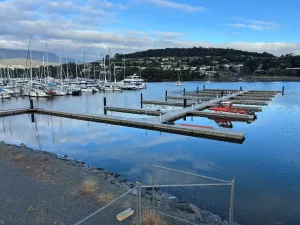 A calm marina in Bellerive with sailboats docked on the left, empty boat slips in the center, and hills with houses in the background under a partly cloudy sky. A gravel path and a closed metal gate are in the foreground.