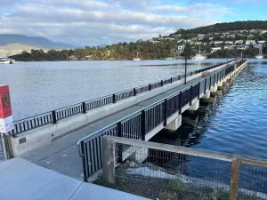 A concrete footbridge with black railings extends over calm water from Bellerive Public Pier toward a tree-lined shore and houses on a hillside under a partly cloudy sky.