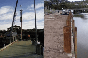Workers are installing large steel piles along a waterfront using cranes and heavy machinery in a land-based piling operation.