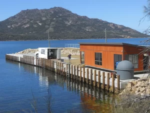 A small house on a dock with a mountain backdrop, surrounded by water and trees.