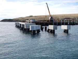 A partially constructed pier at Darlington Jetty extends over the water, with concrete platforms resting on wooden pillars. A crane and construction equipment are visible on the shore in the background.