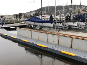Docked sailing boats covered with blue tarps sit on trailers at the Derwent Sailing Squadron Boat Ramp and Hard Stand, with masts visible in the background. The dock has yellow bumpers, and the area reflects a cloudy sky.