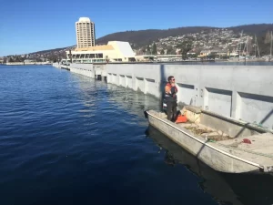 A person in a high-visibility vest stands on a small metal boat near the Derwent Sailing Squadron Break-wall, with modern buildings and houses dotting the hillside in the background under a clear blue sky.