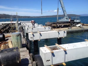 A worker stands on a pier aligning large concrete beams as part of the Dover Jetty Reconstruction, while a crane on a floating platform assists with construction over the blue water on a sunny day.