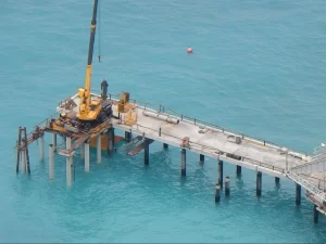 A yellow crane operates on the concrete pier of the Flying Fish Cove Jetty Extension, stretching over clear blue water. A worker stands nearby, with construction materials scattered on the platform and a red buoy floating in the distance.
