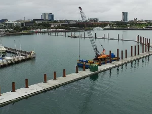 Workers handling and driving large steel piles into the seabed as part of the yacht club's breakwater reinforcement project
