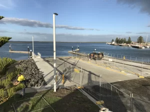 A concrete boat ramp leads into a calm body of water, bordered by rocks and fencing. Several docks extend into the water, with a few boats and a person in a yellow shirt working near the ramp.