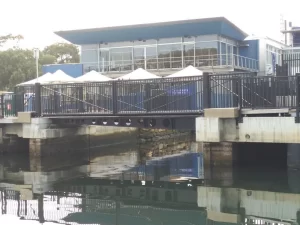 A modern two-story building with large windows and white canopy tents on the balcony overlooks Kangaroo Bay, situated next to a black metal fence and overhanging a body of water reflecting the structure.