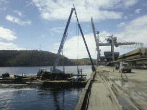 A crane mounted on a barge is installing a mooring dolphin structure beside an industrial dock along a waterfront.