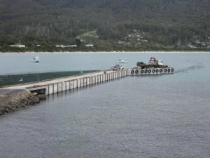 A concrete pier extends into the water at Pirates Bay Jetty, with a vehicle and equipment at the end; boats are anchored nearby and forested hills are in the background.