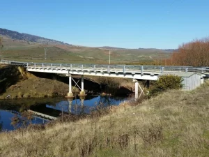 The Rotherwood Bridge Re-Deck features a metal bridge with guardrails spanning over a calm river in a rural landscape, surrounded by grassy hills and distant mountains under a clear blue sky.