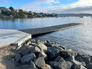 A floating dock extends out into calm water from a rocky shore in Sandy Bay, with houses and trees visible on the opposite bank under a partly cloudy sky.