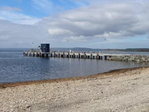 A long pier extends over calm water from a rocky, pebbled beach beneath a mostly cloudy sky, with hills in the distance—capturing the tranquil setting near the Strahan Aquaculture Hub.