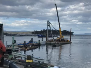A crane mounted on a floating platform lifts equipment near the Tassal Meads Mooring Dolphins on a cloudy day, with boats, docks, and a forested shoreline visible in the background.
