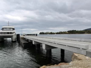 A modern concrete jetty from the Woodbridge Jetty Replacement project extends over calm water with a catamaran docked on the left. A cloudy sky and distant shoreline with trees are visible in the background.